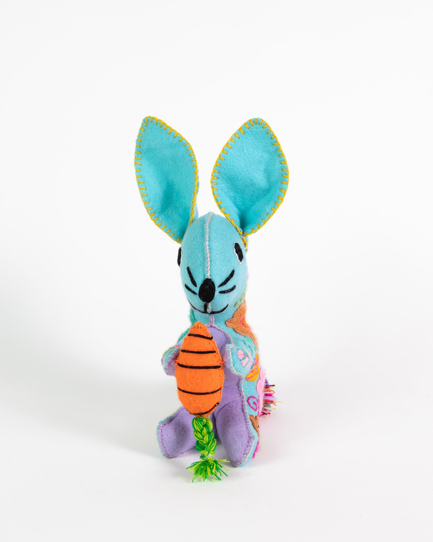 A colorful handmade plush bunny toy holding an orange striped carrot with green tinsel, photographed against a plain white background.