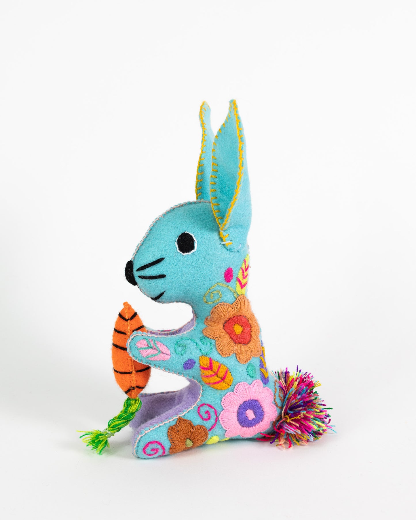 A handmade felt bunny plush toy in light blue, decorated with colorful embroidered flowers, holding a small orange carrot, on a plain white background.