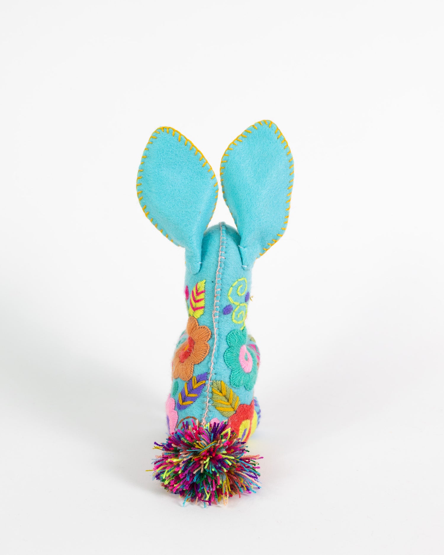 A turquoise blue felt bunny with long upright ears, colorful embroidered floral details, and a multicolor tinsel pom-pom at the bottom, photographed on a clean white background.